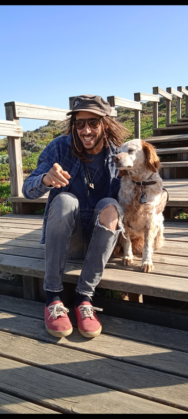 A joyful man in sunglasses and a cap sits on wooden steps with his happy dog. Sunny day, outdoor setting, lots of greenery, relaxed atmosphere, genuine smiles.