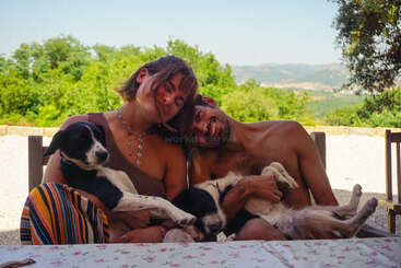 A smiling couple sits at an outdoor table, each holding a relaxed dog. Trees and mountains create a peaceful, sunny background. The scene radiates warmth and happiness.