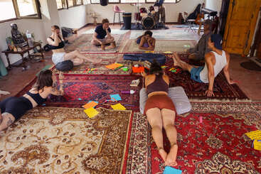 A group of people relax and interact in a cozy room with colorful rugs, art supplies, and musical instruments, creating a laid-back, creative, and communal atmosphere.