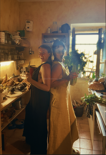 Two women stand back-to-back in a cozy, sunlit kitchen, smiling playfully; one holds a knife, the other celery, surrounded by fresh vegetables and warm light.