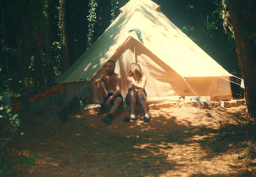 A man and woman sit outside a large beige tent in a forest, enjoying sunlight and nature, surrounded by trees and camping gear, looking relaxed and happy.