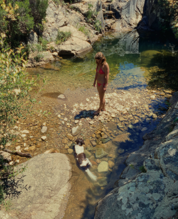 A woman in a pink bikini stands on river rocks, while a black and white dog wades nearby in the shallow, clear water surrounded by rocky cliffs.