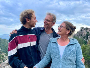 Three people stand outdoors, smiling and looking at each other warmly. They wear casual jackets. Behind them are green hills, rocky cliffs, and a cloudy blue sky.