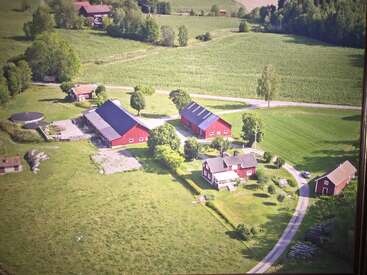 This aerial image shows a rural farm with red barn buildings, a farmhouse, green fields, scattered trees, a winding road, and a peaceful countryside landscape.