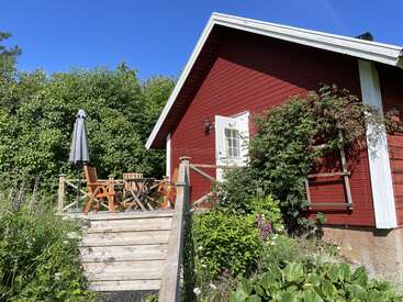 A charming red cottage with white trim sits surrounded by lush greenery. A wooden deck with table, chairs, and an umbrella invites relaxation under clear blue skies.