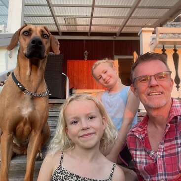 A man, two young girls, and a large brown dog sit on wooden steps outside a house, smiling and enjoying a sunny, relaxing moment together.