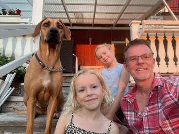Un hombre, dos niñas y un gran perro marrón están sentados en unos escalones de madera en el exterior de una casa, sonriendo y disfrutando juntos de un momento soleado y relajante.