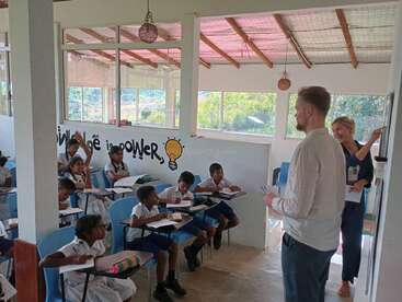 Une salle de classe où des élèves en uniforme écoutent attentivement deux enseignants. Sur le mur, on peut lire « La connaissance, c'est le pouvoir », accompagné d'une illustration représentant une ampoule. Une salle lumineuse et ouverte, un apprentissage stimulant.