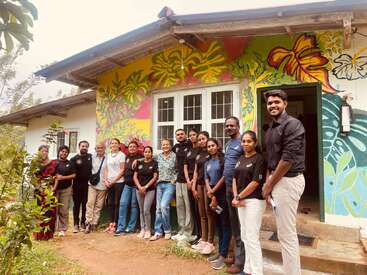 Un groupe de personnes sourit devant un bâtiment coloré orné de peintures murales. L'arrière-plan est décoré de feuilles et de fleurs tropicales, suggérant une atmosphère joyeuse et communautaire.