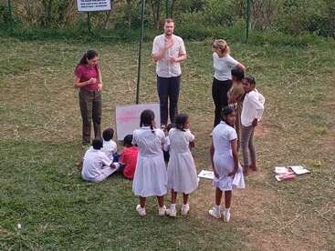 Un groupe d'enfants et d'adultes suit un cours en plein air sur l'herbe, avec un tableau blanc, des cahiers et des discussions. Certains élèves portent des uniformes blancs.