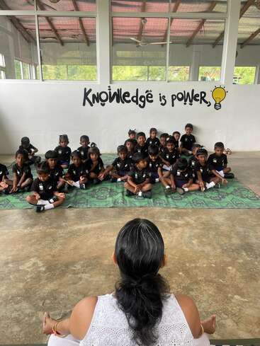 Un groupe de jeunes enfants est assis en tailleur sur des tapis, face à un enseignant. Derrière eux, sur le mur, on peut lire « La connaissance, c'est le pouvoir », accompagné d'une illustration représentant une ampoule jaune.