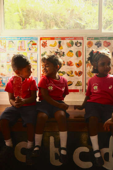 Trois jeunes enfants vêtus d'uniformes assortis sont assis ensemble sur un banc, l'un d'eux dégustant une glace. Des tableaux colorés représentant des fruits et la lumière du soleil créent une atmosphère joyeuse dans la salle de classe.