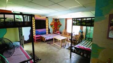 Este quarto colorido e animado tem beliches, camas de solteiro, cadeiras de vime, um lustre, paredes pintadas, um ventilador e uma mesa de madeira no meio, sobre um tapete.