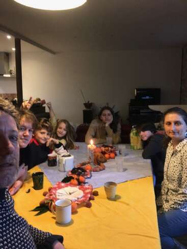 A family gathers around a festive table with mugs, fruit, and a candle centerpiece. Everyone looks toward the camera, sharing a warm, cozy, indoor moment together.