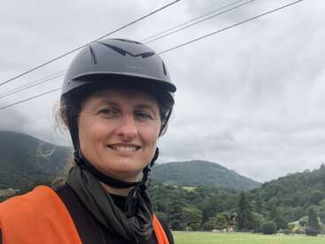 La imagen muestra a una mujer con casco negro y chaleco de seguridad naranja, sobre un fondo de árboles y colinas bajo un cielo nublado en el que se ven líneas de alta tensión.