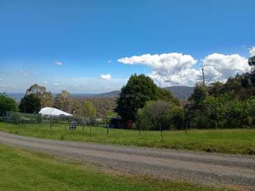 A scenic rural landscape featuring a gravel road, green grassy fields, scattered trees, farm buildings, distant hills, and a bright blue sky with fluffy clouds.