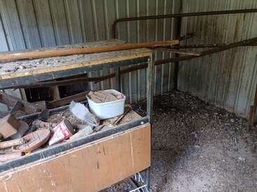 This image shows an old, dusty, abandoned room with a metal wall, cluttered wooden table, scattered debris, and signs of neglect and decay throughout the space.