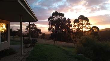 A peaceful countryside scene at sunset, with a house, potted plants, grassy yard, and tall silhouetted trees against a colorful sky filled with soft clouds.