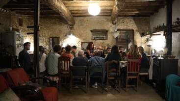 Un grupo de personas se reúne alrededor de una gran mesa en una sala rústica y acogedora, con paredes de piedra y vigas de madera, compartiendo una comida bajo una cálida iluminación.