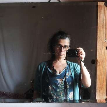 Une femme à lunettes prend un selfie devant un miroir, un appareil photo à la main. Vêtue d'une chemise à fleurs bleue et d'un cardigan, elle se tient debout dans une pièce rustique et faiblement éclairée.