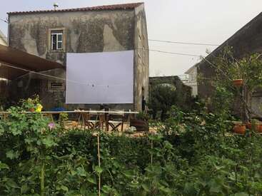 Una casa antigua con una fachada desgastada por el paso del tiempo, un jardín en la parte delantera, mesas y sillas al aire libre, y una gran pantalla blanca colgada para una noche de cine al aire libre o cualquier otro evento.