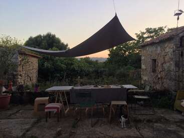This image shows a rustic outdoor dining area between old stone buildings at sunset, with assorted chairs, a table, a sunshade, greenery, and a small cat.