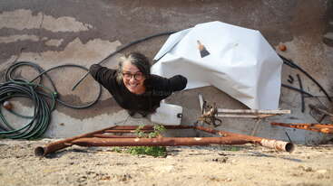 A cheerful person with glasses looks up and smiles at the camera while standing near a wall, surrounded by hoses, metal pipes, and tools.