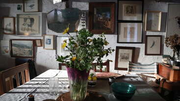 A sunlit dining room features a table with a vibrant bouquet in a vase, surrounded by eclectic chairs. The wall is decorated with framed artwork and photos.