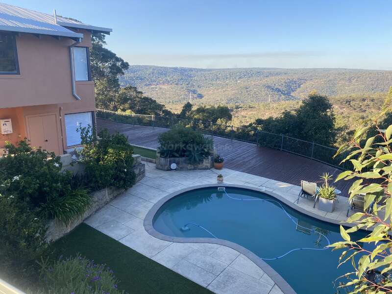 A serene hillside home with a curved swimming pool, lush garden, wooden deck, and stunning panoramic views of green valleys and distant rolling mountains under a clear sky.