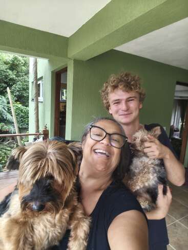 Uma mulher feliz e um rapaz sorriem para uma selfie na varanda, cada um segurando um cachorro fofinho, curtindo um momento alegre juntos ao ar livre.