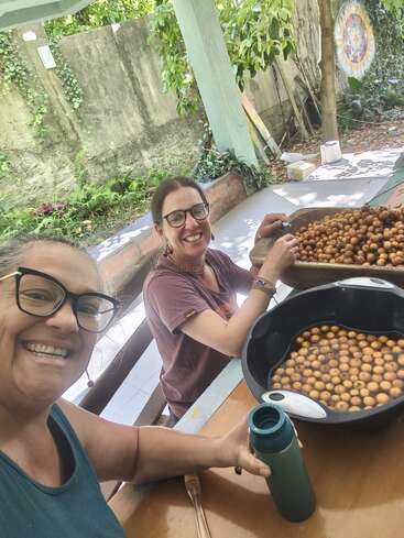 Duas mulheres sorrindo ao ar livre, sentadas à mesa com tigelas de pequenas frutas redondas. Uma delas segura uma garrafa. Uma vegetação exuberante e um mural colorido decoram o fundo.