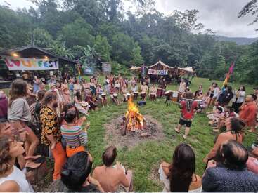 Un grupo numeroso se reúne alrededor de una hoguera en un animado festival al aire libre. La gente toca tambores, aplaude y disfruta de la música, rodeada de coloridas tiendas de campaña y exuberante vegetación.