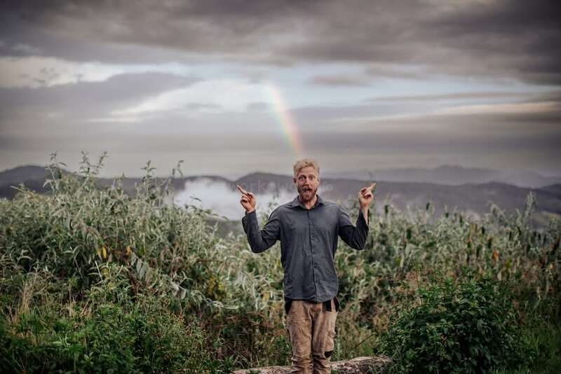 Un hombre, de pie frente a la hierba alta y las montañas, señala entusiasmado un vibrante arco iris en el cielo nublado a sus espaldas, creando un ambiente de alegría.