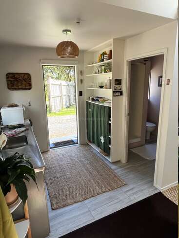 Das ist eine kleine, aufgeräumte Küche mit offenen Regalen, grünen Vorhängen, einem gewebten Teppich, einer Edelstahltheke, einer Pflanze und einer sichtbaren Badezimmertür.