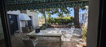 Un patio paisible avec des chaises en fer forgé blanc et une table sous un auvent à motifs verts. Des documents et un ordinateur portable sont posés sur la table à côté d'un jardin.