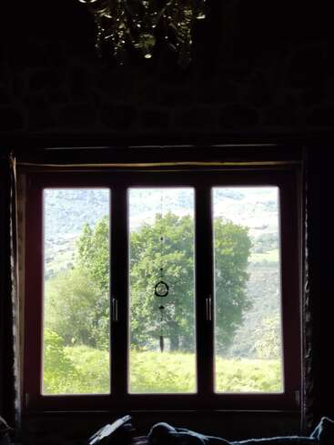 Une grande fenêtre encadre une vue panoramique sur des arbres verdoyants et des collines lointaines, avec un capteur de rêves suspendu au centre et un lustre au-dessus.