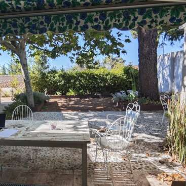 Ein friedlicher Innenhof mit weißen schmiedeeisernen Stühlen und einem Tisch unter einer grün gemusterten Markise. Auf dem Tisch neben dem Garten liegen Papiere und ein Laptop.