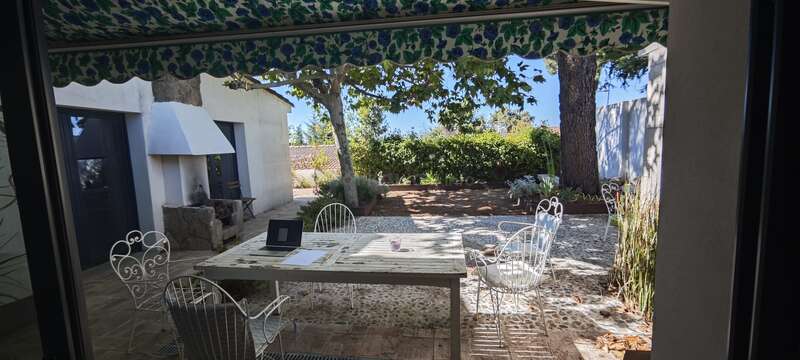 Ein friedlicher Innenhof mit weißen schmiedeeisernen Stühlen und einem Tisch unter einer grün gemusterten Markise. Auf dem Tisch neben dem Garten liegen Papiere und ein Laptop.