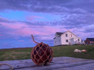 Uma boia de vidro rústica enrolada em corda está sobre uma mesa de madeira desgastada pelo tempo. Ao fundo, uma casa branca está sob um lindo céu roxo e rosa.