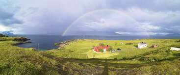 Um arco-íris vibrante se estende sobre uma paisagem verde exuberante com casas espalhadas perto de um litoral rochoso, sob um céu nublado e dramático, ao lado de um oceano azul e calmo.