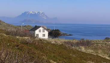 A imagem retrata uma cena costeira serena com uma casa branca com um telhado escuro, tendo como pano de fundo uma montanha majestosa e um vasto corpo de água.