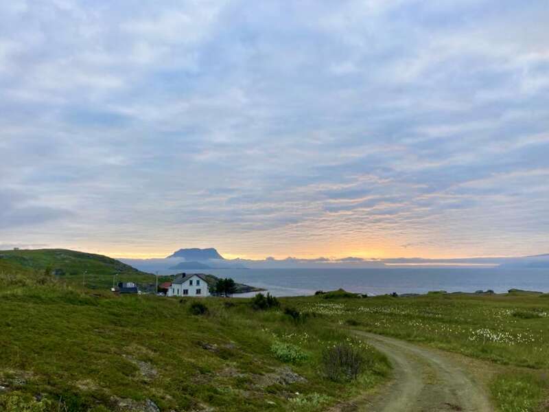 Uma cena litorânea tranquila mostra uma casa branca solitária, campos verdes, uma estrada de terra sinuosa, águas calmas do oceano, montanhas distantes e um céu suavemente brilhante ao pôr do sol.