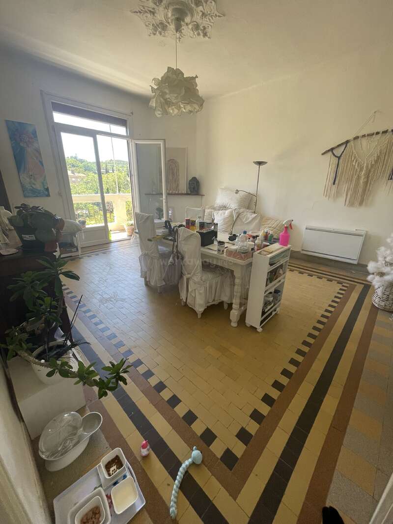 Uma sala aconchegante e ensolarada com azulejos antigos, móveis brancos, janelas abertas, plantas, arte nas paredes, uma estação para alimentar animais de estimação e vários itens espalhados sobre a mesa.