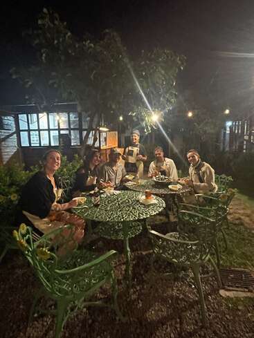 Six personnes prennent un repas nocturne en plein air autour d'une table en métal vert, entourée de plantes et de lumières, créant ainsi une atmosphère chaleureuse et détendue dans le jardin.