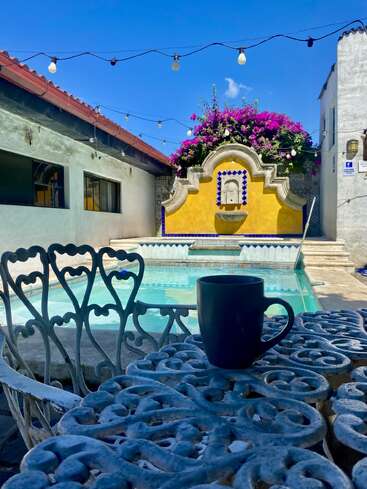 Une tasse à café noire est posée sur une table en métal bleu orné, à côté d'une piscine, avec des guirlandes lumineuses, des fleurs colorées et une fontaine jaune en arrière-plan.
