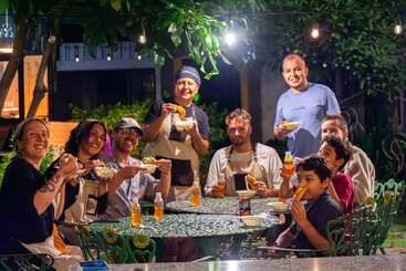 Un alegre grupo de personas, entre las que hay adultos y niños, se sienta alrededor de una mesa al aire libre por la noche, compartiendo comida, bebida y risas bajo unas guirnaldas de luces.