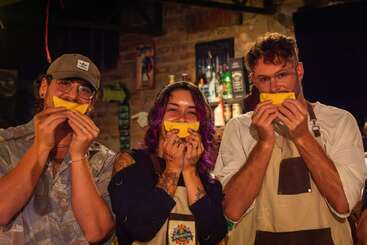 Tres personas están de pie, una al lado de la otra, en un acogedor bar, cada una con comida amarilla con forma de sonrisa en la mano, y parecen felices y juguetonas juntas.
