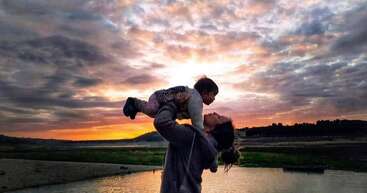 A woman holds a child aloft in front of a sunset over a body of water, with a cloudy sky above.