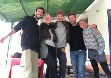 Five smiling people, arm in arm, pose cheerfully under a green roof. They are casually dressed, radiating warmth, friendship, and happiness in a relaxed setting.