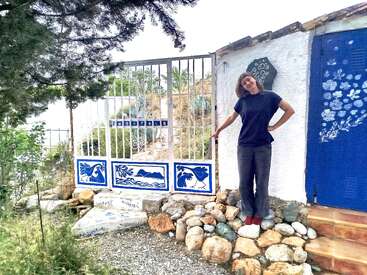 A woman stands smiling beside a gate decorated with blue and white art. The wall and door behind her are painted white and vibrant blue with flowers.